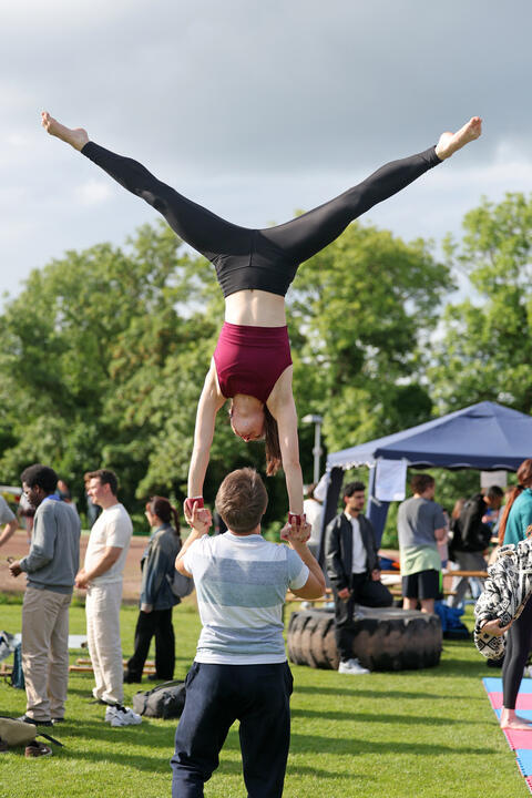 Zwei Personen zeigen gemeinsam eine akrobatische Handstandfigur auf einer Wiese bei einer Veranstaltung.