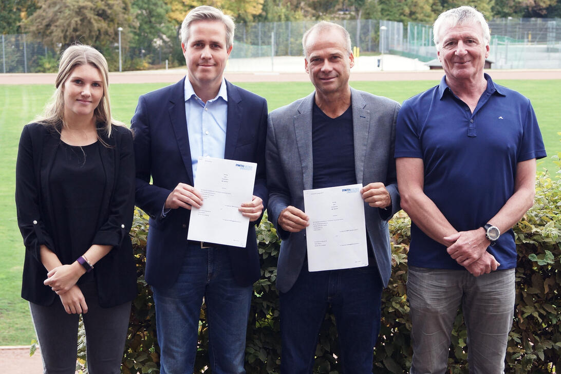 eine Frau drei Männer vor einem Sportpatz mit einem Vertrag in der Hand